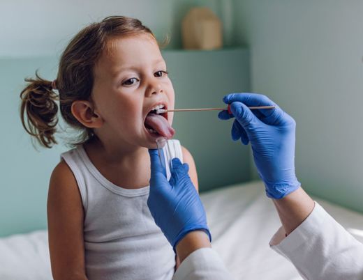 A child with pigtails sits on the table in a doctor’s office. A clinician wearing blue gloves swabs the inside of the child’s mouth with a long cotton swab.
