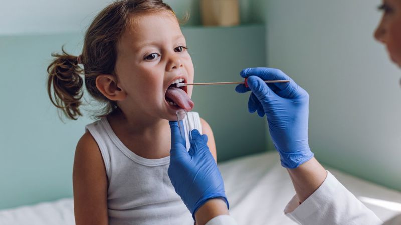 A child with pigtails sits on the table in a doctor’s office. A clinician wearing blue gloves swabs the inside of the child’s mouth with a long cotton swab.