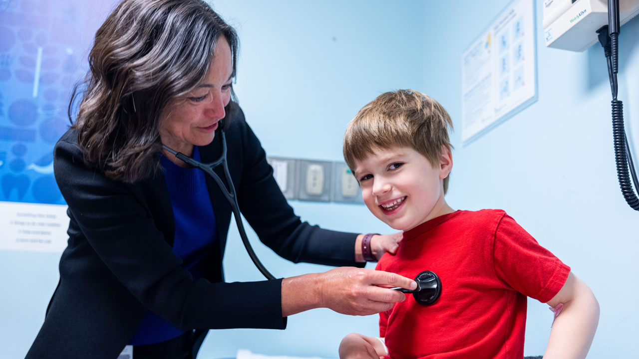 Dr. Shelley Miyamoto uses a stethoscope to listen to the heart of a young child wearing a red shirt.