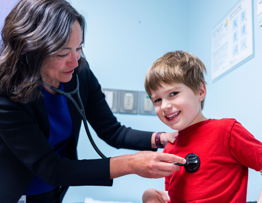 Dr. Shelley Miyamoto uses a stethoscope to listen to the heart of a young child wearing a red shirt.