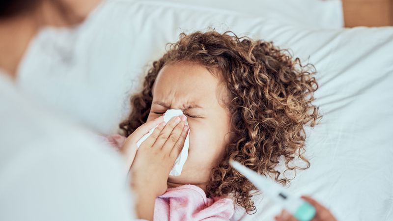 Adult helping child blow their nose while laying in bed with a thermometer 