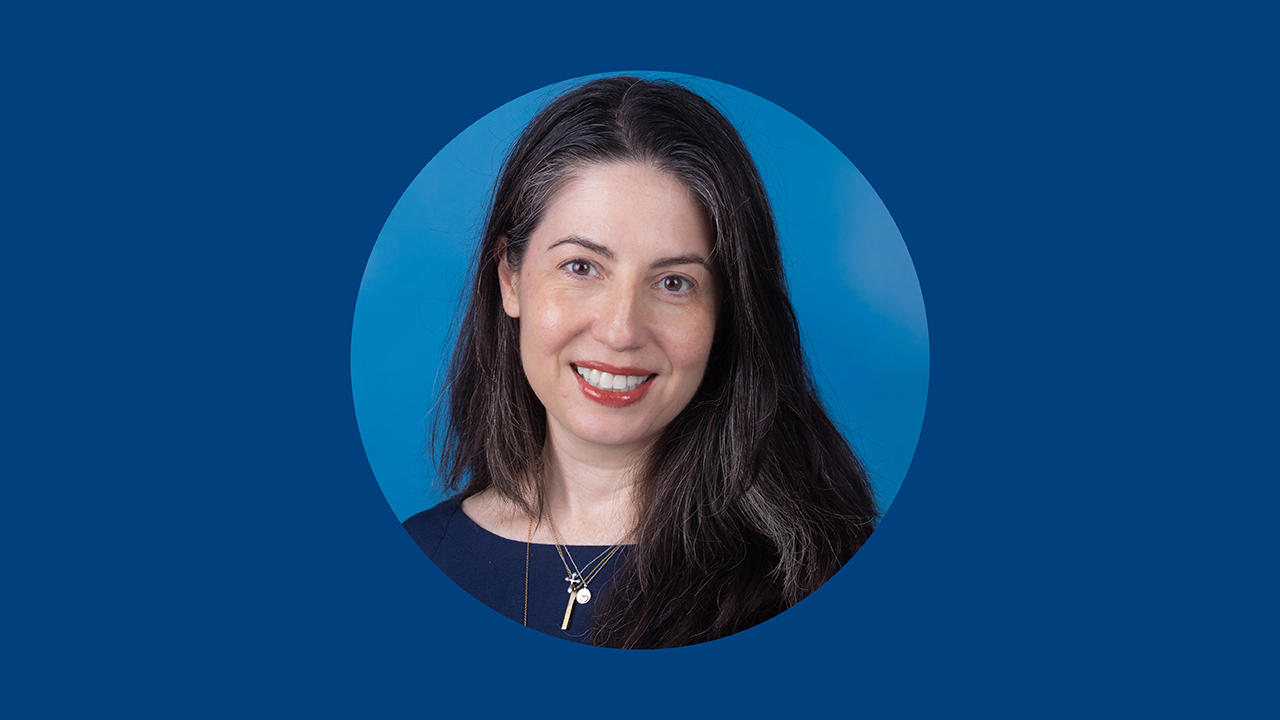 A woman with dark hair wearing multiple necklaces smiles in front of a blue background and in a circular frame.