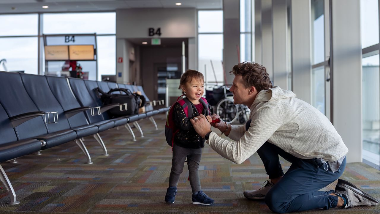 Adult kneeling to adjust a toddler’s backpack while standing in an airport terminal waiting area.