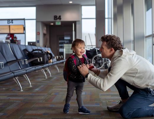 Adult kneeling to adjust a toddler’s backpack while standing in an airport terminal waiting area.