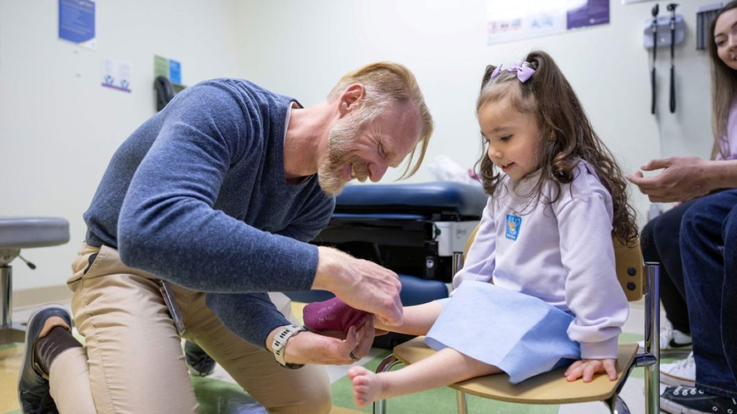 An adult medical provider kneels on the floor in a hospital room and smiles while he puts a sock on the foot of a young child, who sits in a chair while wearing a dress.