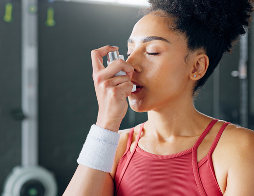 A person in athletic clothing uses an inhaler in a gym setting, holding it up to their mouth with exercise equipment visible in the background.