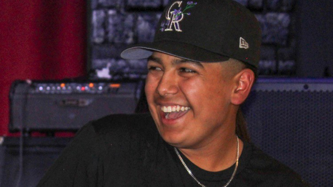 Person wearing a black baseball cap and black shirt smiles broadly while seated indoors, with audio equipment and a red curtain visible in the background.