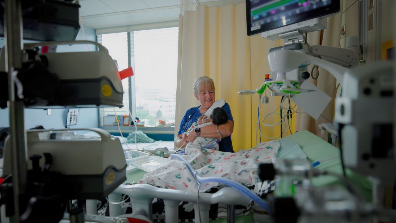 Person holding an infant in a hospital room, surrounded by medical monitors and equipment.