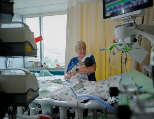 Person holding an infant in a hospital room, surrounded by medical monitors and equipment.