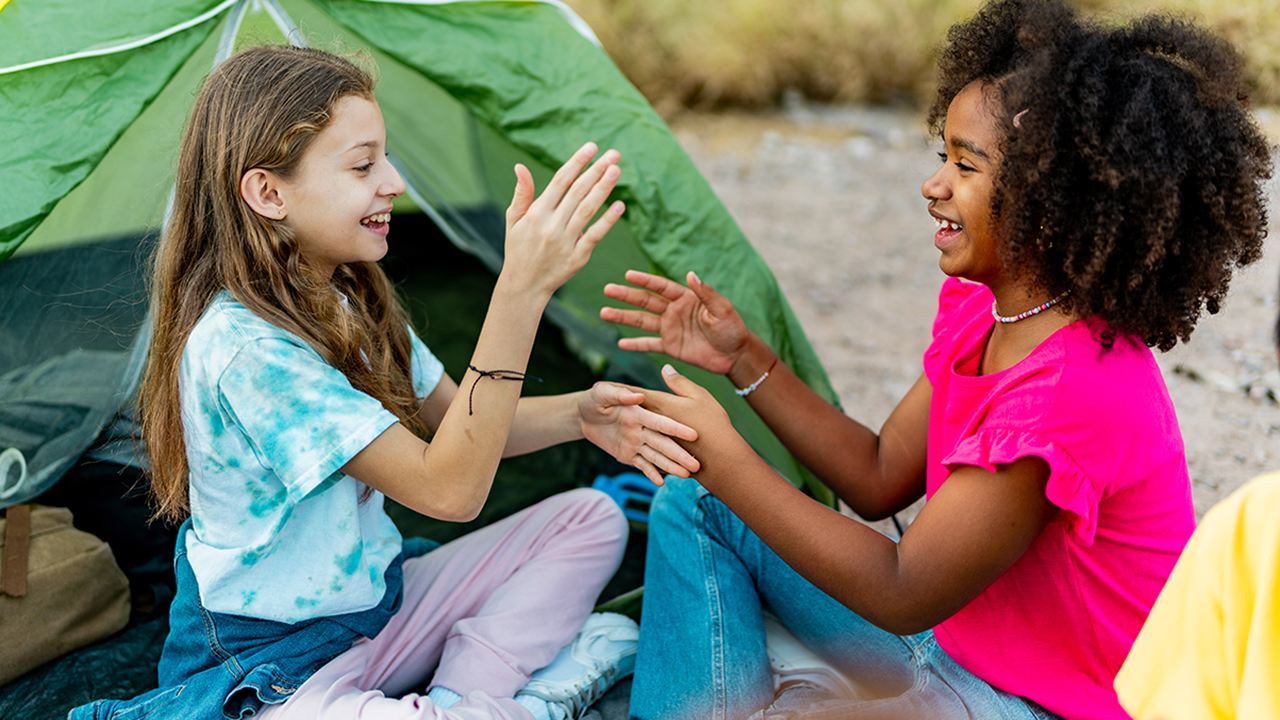 Two children sit on the ground in front of a green camping tent near a rocky stream, facing each other and playing a clapping hand game, with backpacks and outdoor gear nearby.