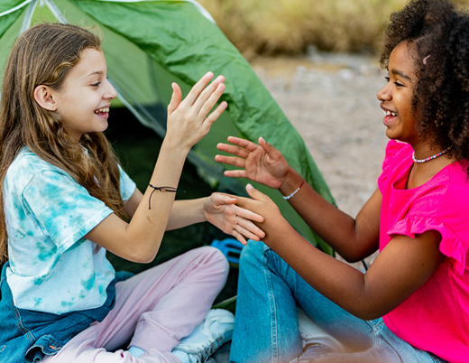 Two children sit on the ground in front of a green camping tent near a rocky stream, facing each other and playing a clapping hand game, with backpacks and outdoor gear nearby.
