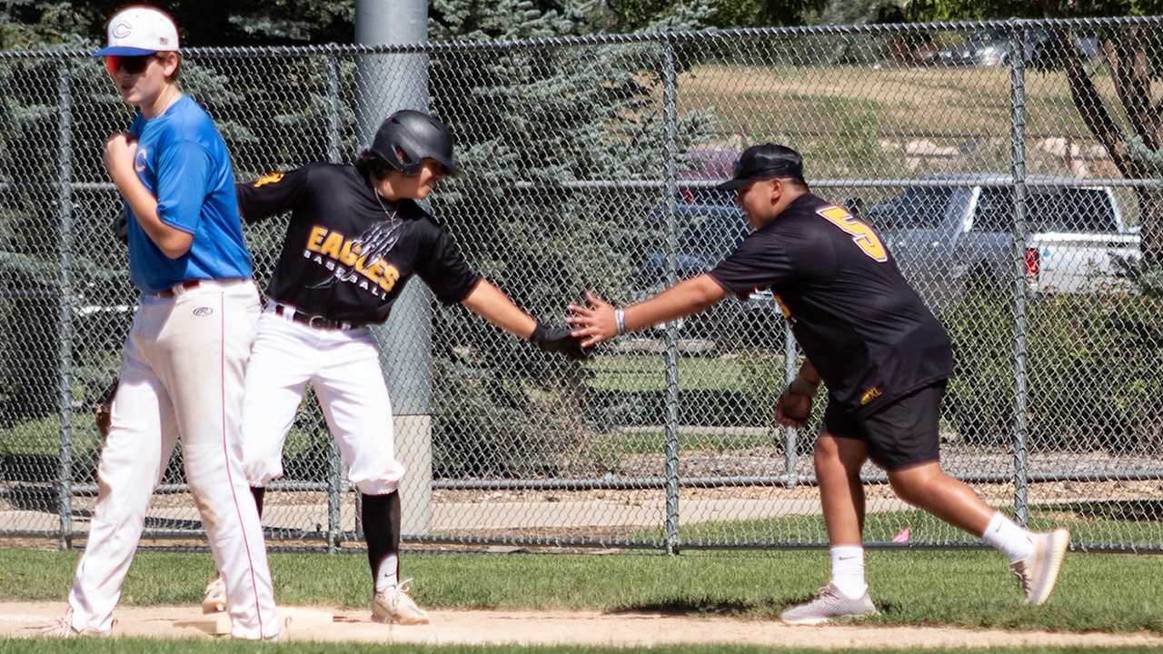Baseball player in a black jersey and batting helmet reaches out to exchange a hand slap with a coach near a base during a game.