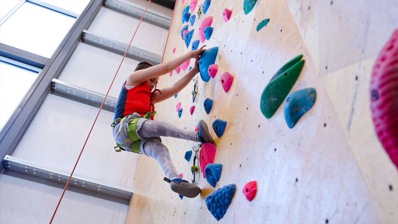 A person in a red top and black shorts is climbing an indoor rock wall fitted with colorful holds.