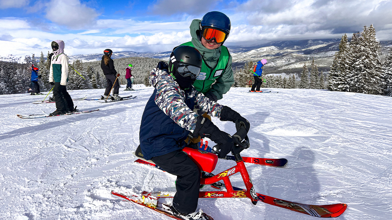 Two individuals using adaptive ski equipment on a snowy mountain, with other skiers in the background under a partly cloudy sky.