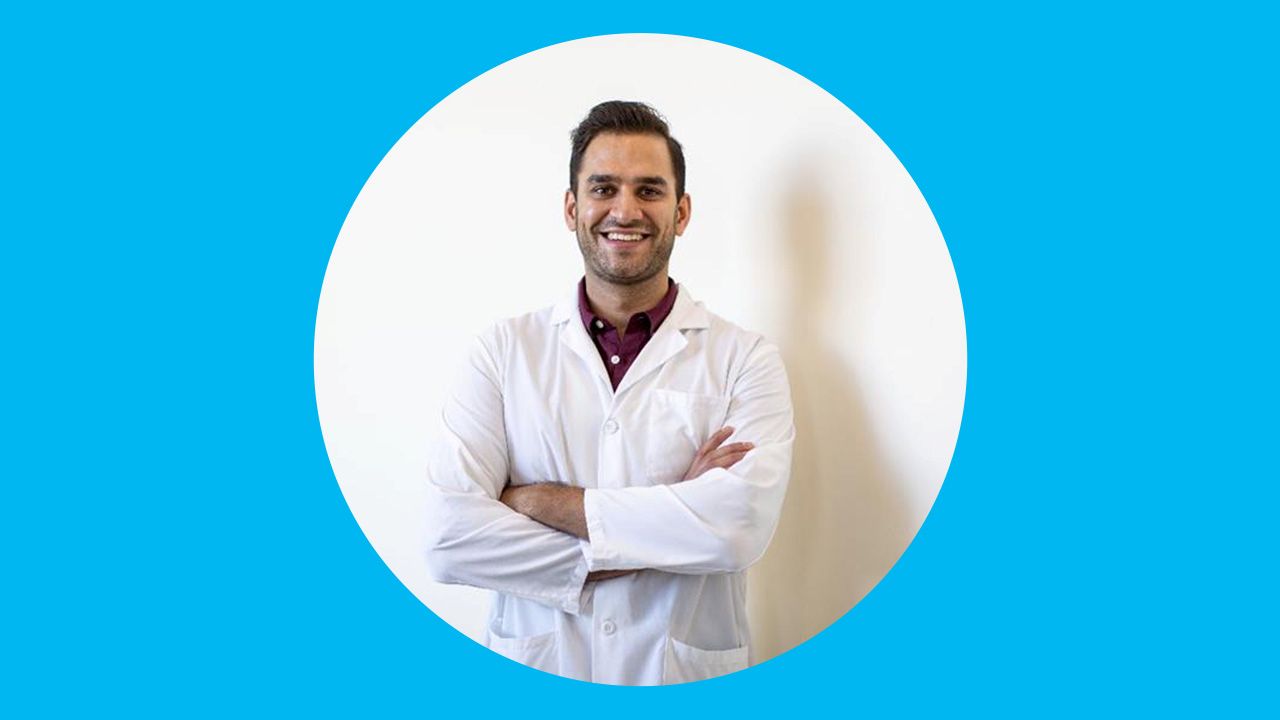 Man stands in a lab coat with his arms crossed in front of a white wall. 