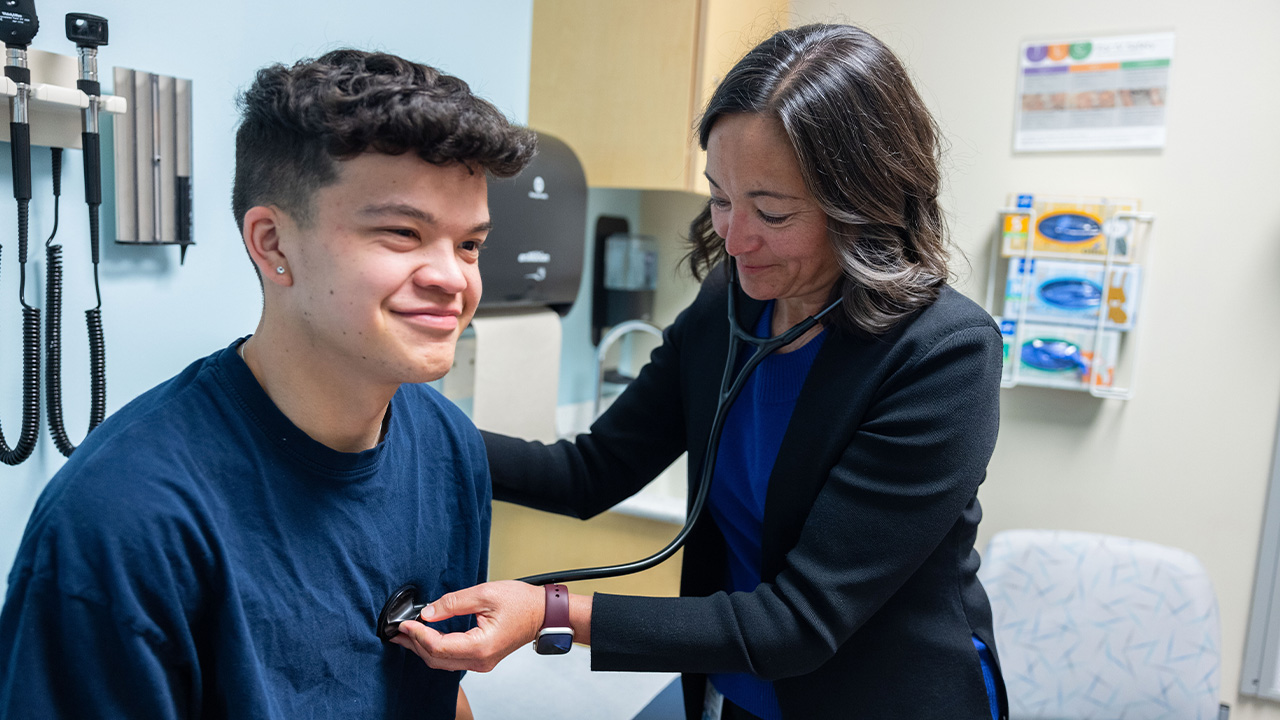 A doctor in a black blazer uses a stethoscope to check the heartbeat of a young teen wearing a navy-blue t-shirt.