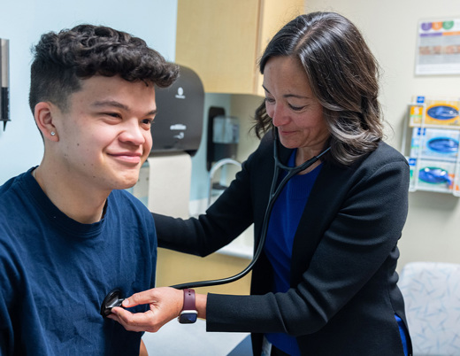 A doctor in a black blazer uses a stethoscope to check the heartbeat of a young teen wearing a navy-blue t-shirt.