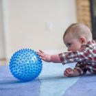 A baby lies on a carpeted floor indoors, reaching one arm toward a round blue textured ball with raised bumps.