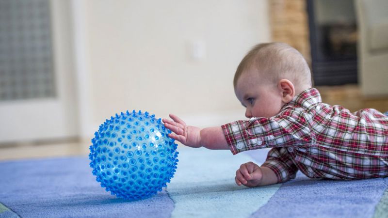A baby lies on a carpeted floor indoors, reaching one arm toward a round blue textured ball with raised bumps.