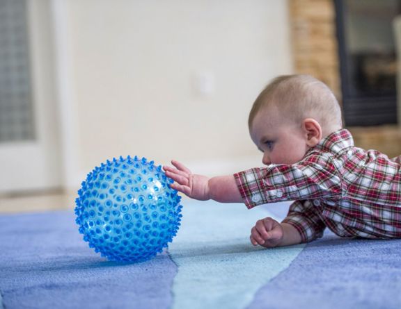 A baby lies on a carpeted floor indoors, reaching one arm toward a round blue textured ball with raised bumps.