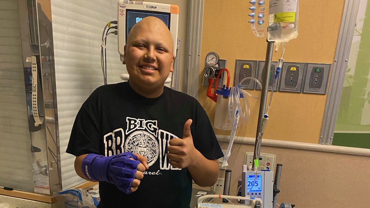 Person standing in a hospital treatment room gives a thumbs-up while wearing a protective hand brace.