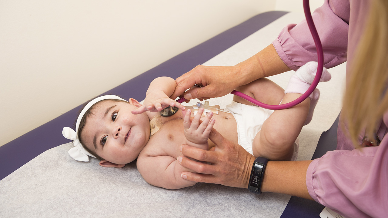 A cardiologist at Children’s Hospital Colorado is examining an infant wearing a diaper and a bow.