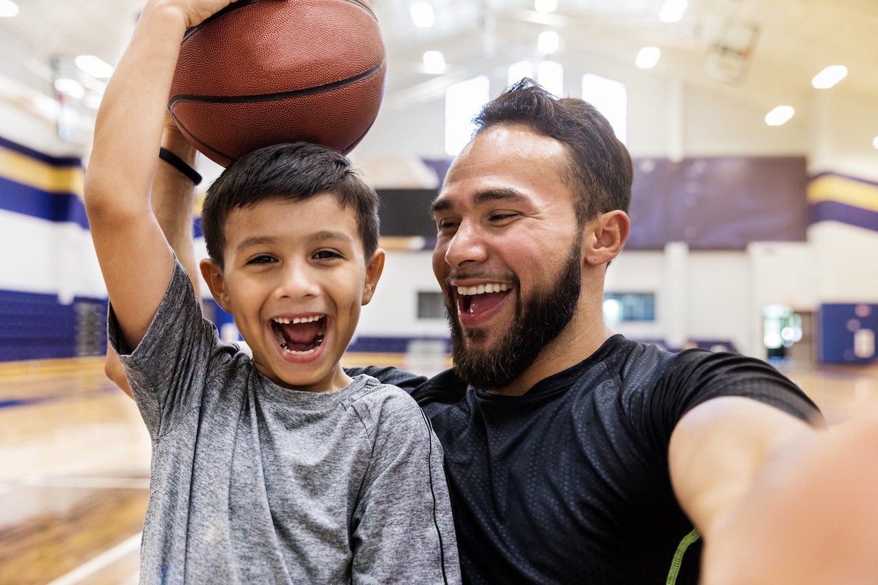 A smiling father stands next to his young son in a gym as the boy lifts a basketball overhead, bothing smiling.