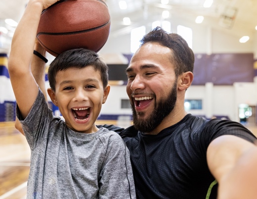A smiling father stands next to his young son in a gym as the boy lifts a basketball overhead, bothing smiling.
