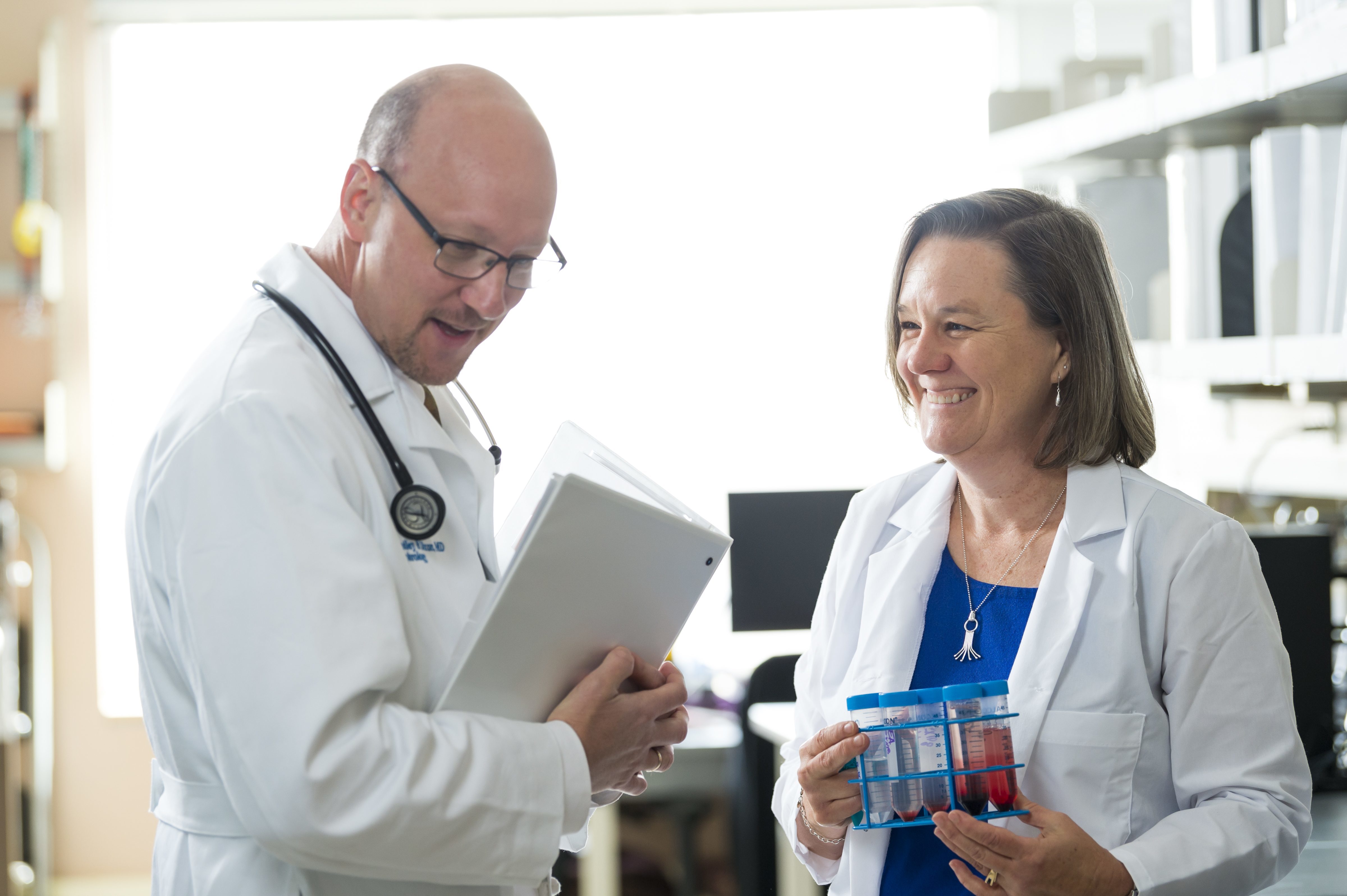 Man in white coat standing with folder and woman smiling with white coat holding test tubes 