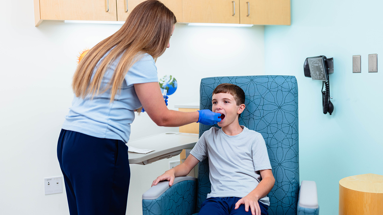A young boy sits in a chair in a hospital room. A nurse with gloves on places something in his mouth. 