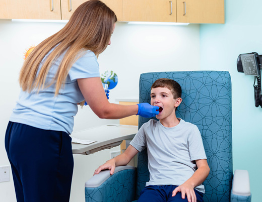 A young boy sits in a chair in a hospital room. A nurse with gloves on places something in his mouth. 