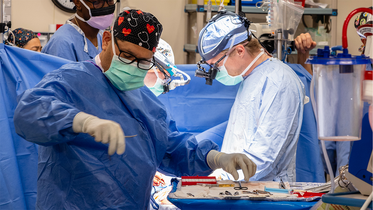 Two surgeons in masks, caps and blue gowns perform a focused operation, handling instruments beside a draped patient in a bright operating room.