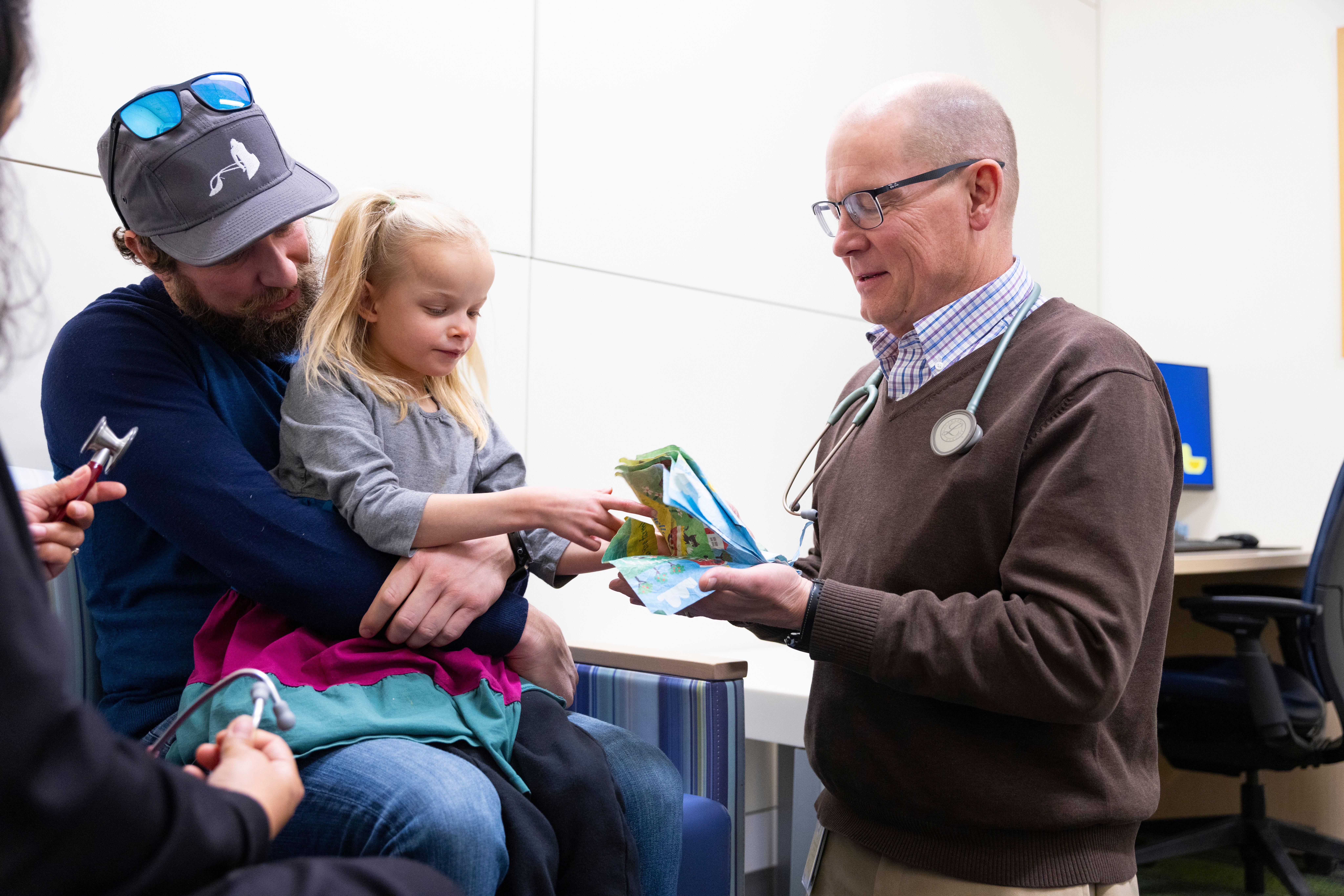 Clinician showing a folded paper to a young child seated on an adult’s lap in a medical exam room.