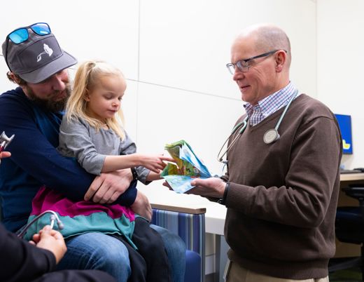 Clinician showing a folded paper to a young child seated on an adult’s lap in a medical exam room.