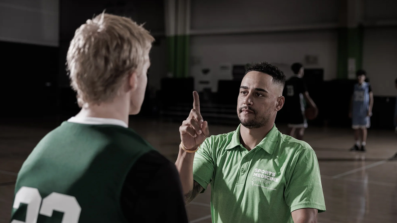 A sports medicine professional in a green polo shirt performs a visual tracking assessment with a young athlete in a gymnasium.
