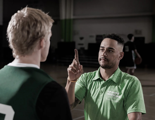A sports medicine professional in a green polo shirt performs a visual tracking assessment with a young athlete in a gymnasium.