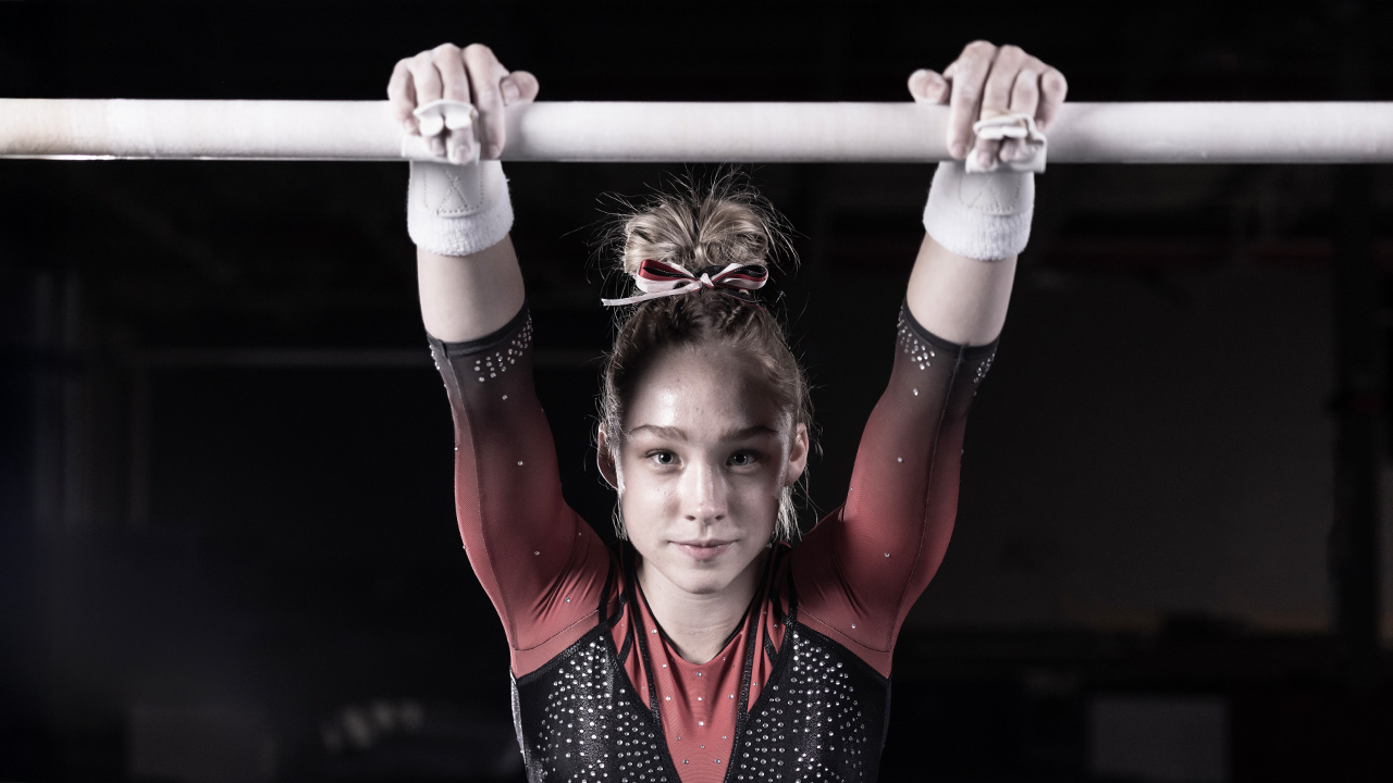 Young gymnast performing on the uneven bars, arms extended overhead in a gymnastics facility.