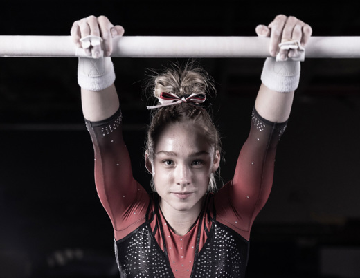 Young gymnast performing on the uneven bars, arms extended overhead in a gymnastics facility.