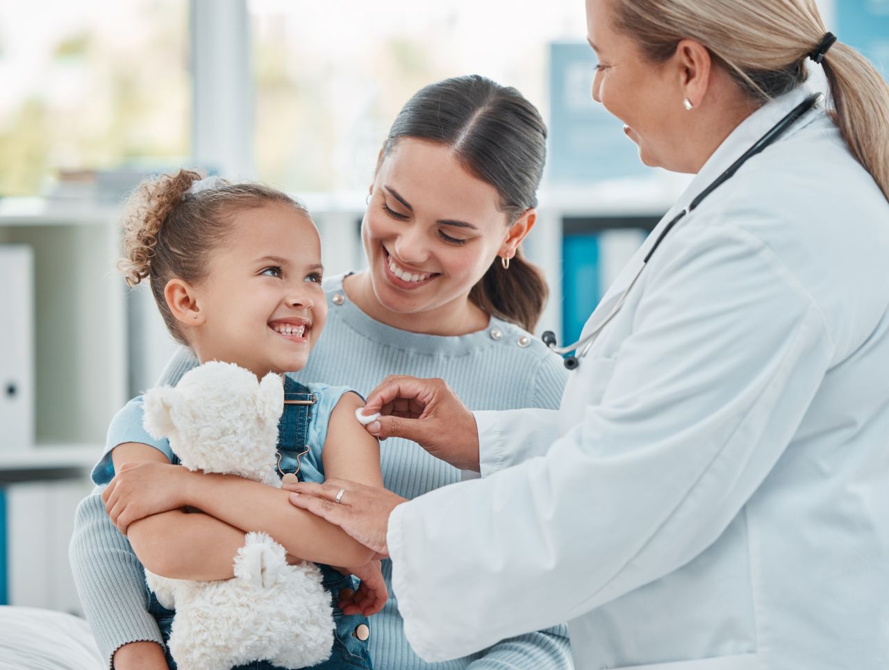 A smiling girl holding a stuffed animal with her mom while a doctor examines her.