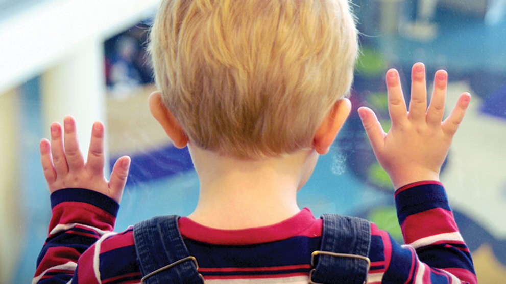 A young blond hair boy wearing a blue and red striped shirt and overalls presses his hands against a window overlooking the Children's Hospital Colorado atrium.