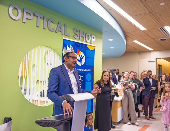 Person wearing a crown standing in front of a crowd of people with the words "Optical Shop" on the wall in the background and a Children's Hospital banner.
