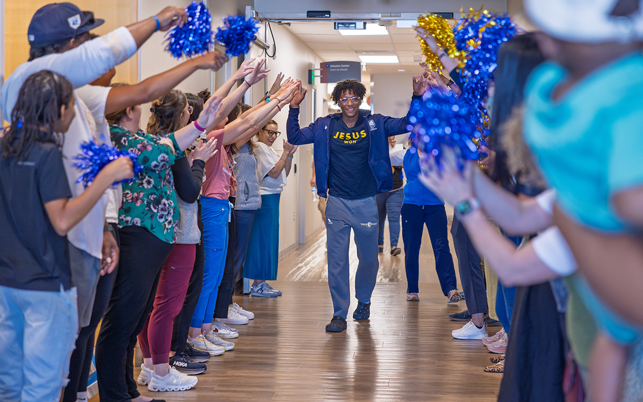 Myles walking down a hallway high fiving family and care team that is lined up against both walls holding blue and yellow pom poms and cheering 