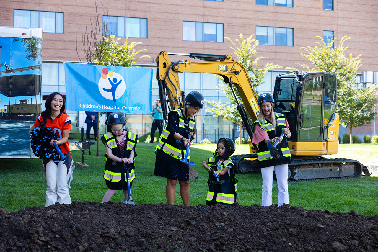 Two kids, two adults and a Denver Broncos cheerleader with shovels digging into a dirt pile on the newest onsite playground with a yellow tractor in the background 