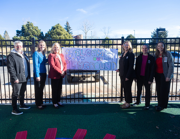 Group of people standing at a playground area in fornt of a sign that reads "Welcome to the Playground" in colorful handwritten writing.