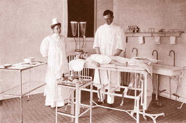 An old photo of the operating room in 1911 with a doctor in a white gown and a nurse in a white gown and white hat standing over a child in a white gown laying on a surgery table; medical supplies and sinks line the back wall