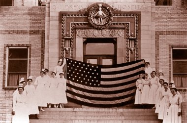 Nurses in white gowns and white hats stand on each side of the steps leading up to the hospital entrance and hold a giant American flag at the top of the stairs