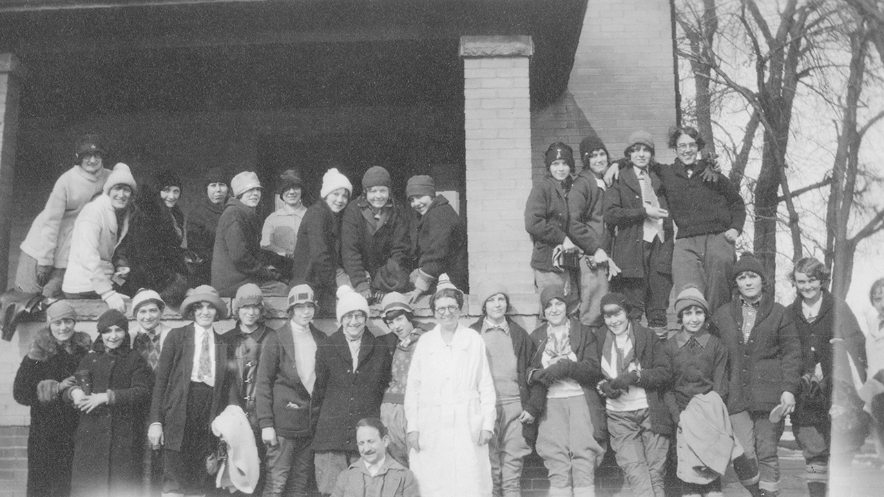 An old photo of about 30 kids gather on the porch - some sitting on the porch wall, others standing in front of it - with a woman in a white gown