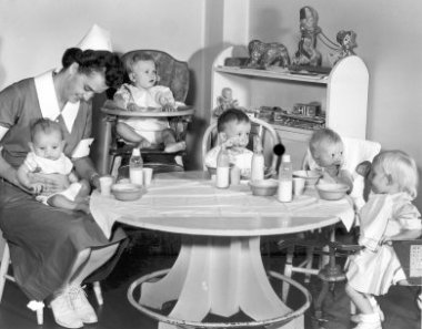 An old photo of a nurse wearing a dress and white hat holding a baby on her lap while another baby sits in a high chair and three toddlers sit around a table eating.