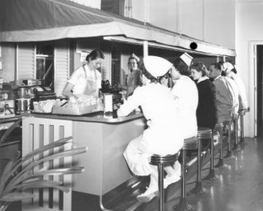 A black and white photo of two women standing under an awning and behind a dining counter serve doctors and nurses who are sitting on stools at a snack bar.