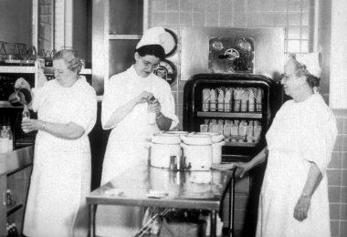 A black and photo of three nurses in white gowns and white hats standing around a cart holding three large canisters in the middle of the room. One is pouring liquid into a bottle, another is putting the cap on, and the third watches the other two.